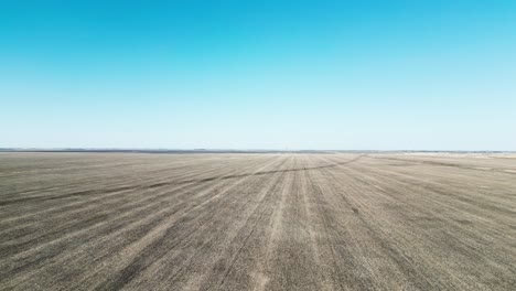 drone floats over barren dirt field; horizon meets cloudless blue sky