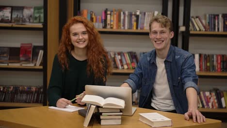 dos jóvenes estudiantes europeos masculinos y femeninos sentados en la mesa con libros y laptop en la biblioteca preparándose para los exámenes