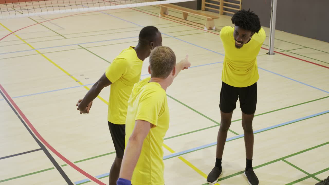 Diverse male team in yellow lifting heads, stretching arms on court by volleyball net warming up