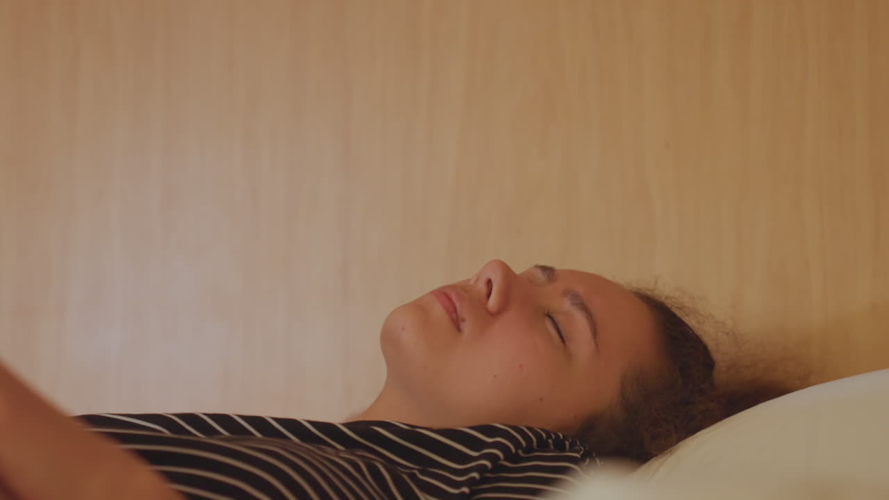 En kvinde i en hotelseng kæmper med søvnløshed og træthed. Hun vrider sig uroligt under dynen, hendes hænder klamrer sig til lagenet, og hendes åndedræt er overfladisk. Et intimt nærbillede viser et anspændt udtryk i lav, varm belysning.