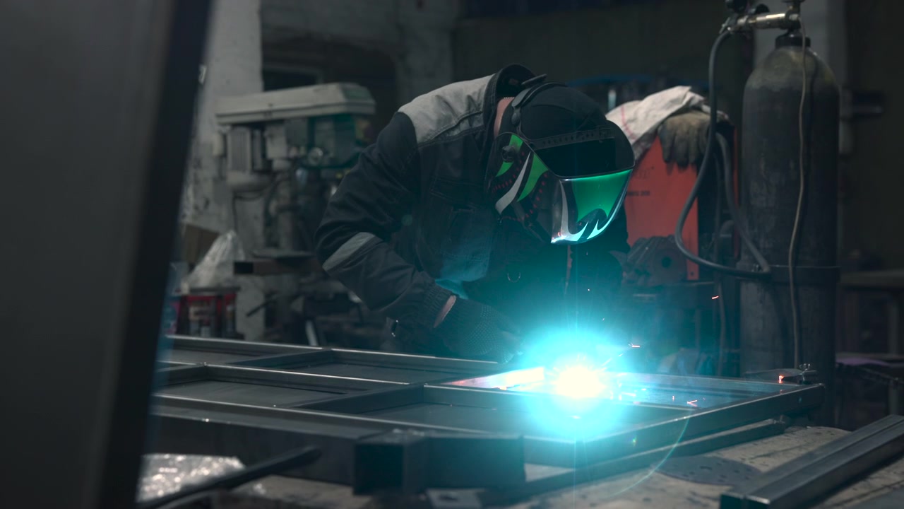 Single worker in protective welding helmet bends over metal fabrication bench inside dim industrial workshop, with bright welding arc, sparks, cluttered tools, pipes, cables, drill press, concrete