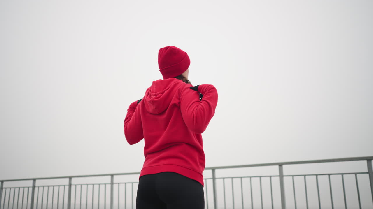 은 후디, 검은 장갑 및 빨간 베니를 입은 여성의 뒷면으로 왼쪽으로 팔과 머리를 돌리는 야외 설정, 철 레일링 및 먼 안개 분위기
