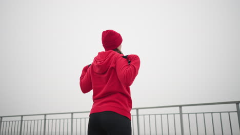 vista trasera de una mujer con capucha roja, guantes negros y gorra roja girando el brazo y la cabeza hacia la izquierda en un entorno al aire libre, con barandilla de hierro y atmósfera de niebla distante