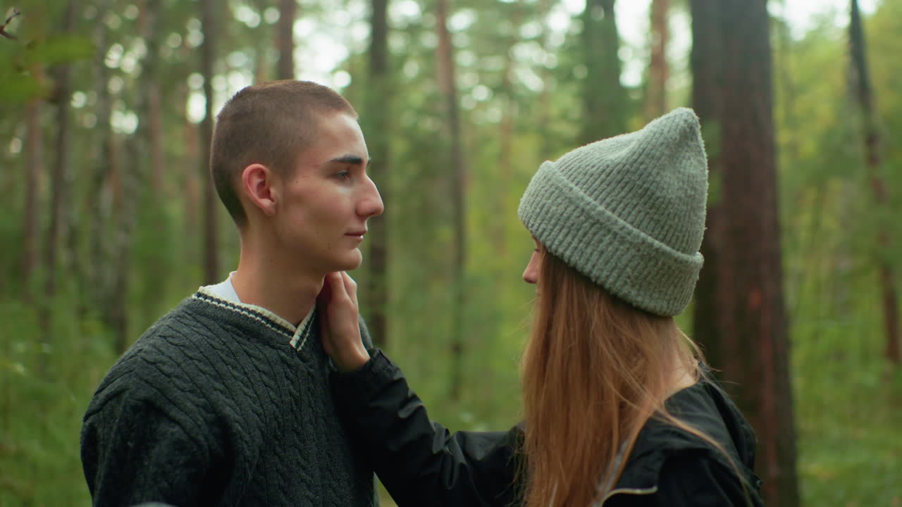 Boy gazes at female companion in forest as she gently touches his cheek during quiet moment, pulling down beanie he used to cover her face playfully, surrounded by tall trees and green background