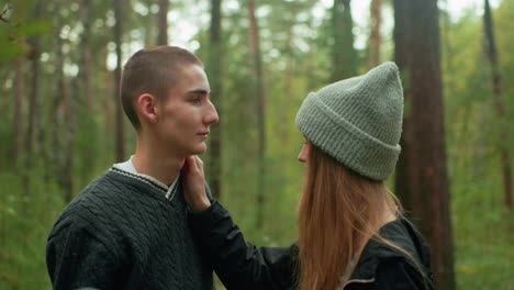boy gazes at female companion in forest as she gently touches his cheek during quiet moment, pulling down beanie he used to cover her face playfully, surrounded by tall trees and green background