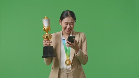 asian business woman in a suit with a gold medal and trophy looking at smartphone then screaming goal celebrating on green screen background in the studio