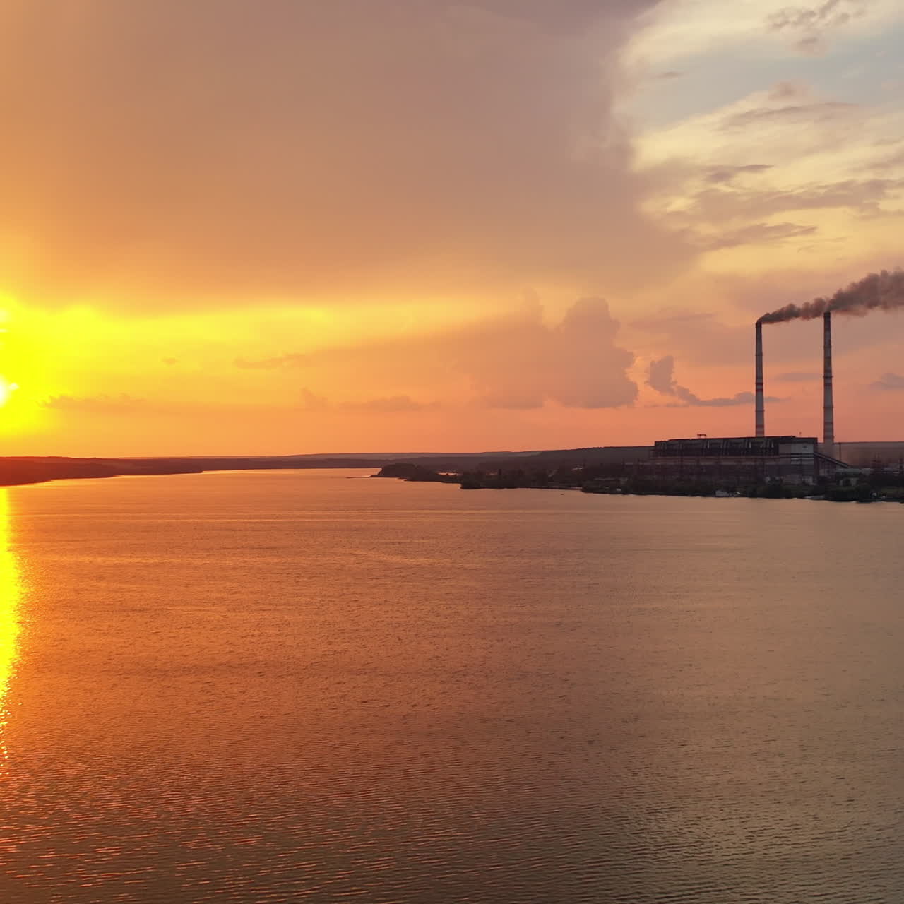 Flying over the river into the big factory at sunset. Two big pipes with white smoke from the power plant near the water background. Motion camera forward. Aerial view.