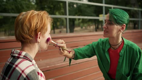 A-girl-with-short-green-hair-in-a-green-shirt-makes-makeup-for-her-friend-with-short-orange-hair-in-a-checkered-shirt-on-brown-benches-in-the-park.-Lesbian-couple-of-LGBT-girls-in-bright-clothes-and-bright-appearance-in-the-park-doing-make-up