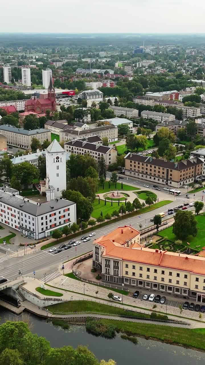 Vertical aerial shot captures Jelgava’s central district, dominated by the striking white Holy Trinity Church Tower and its clock, with historic quarters, orange-roofed buildings, and green parks