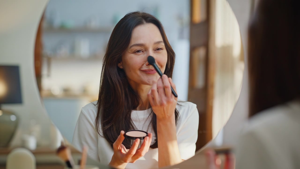 Woman applying face powder using brush looking mirror at beauty routine closeup
