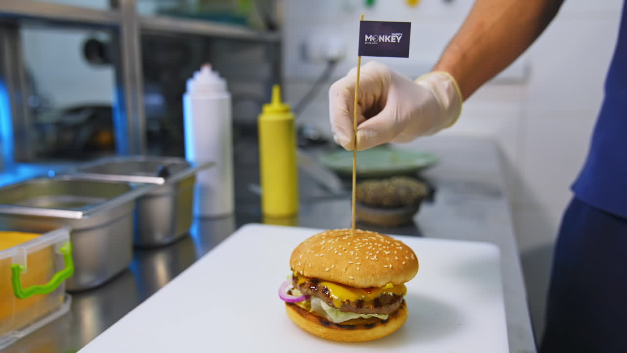 Cook is finishing burger with wooden stick. Stock footage. Preparing burger. Cook hands preparing and making hamburger. Rasty unhealthy dish.