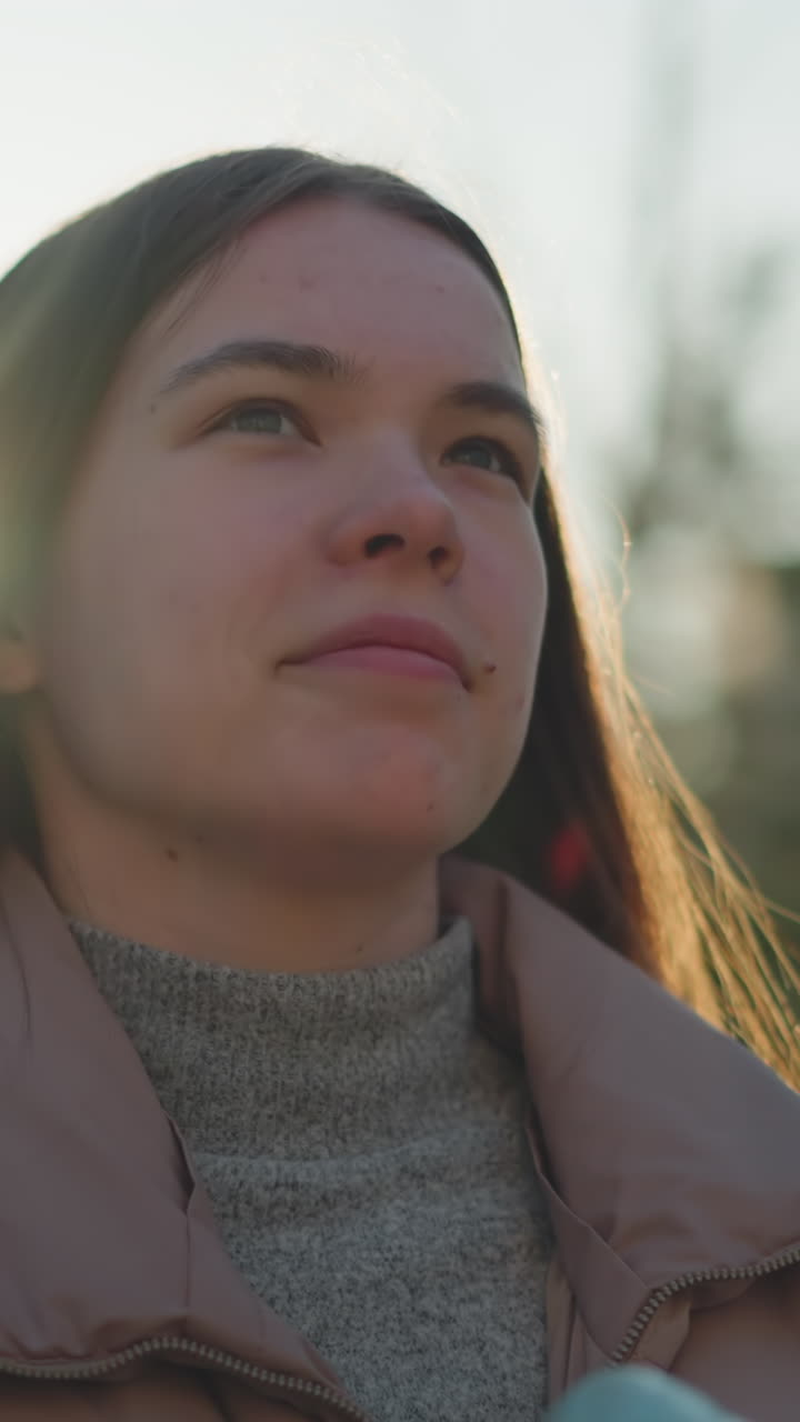 un primer plano de una mujer joven con una chaqueta de melocotón y un suéter de cuello gordo gris, de pie en un parque iluminado por el sol. ella está sosteniendo un par de patines y mirando hacia arriba con una expresión reflexiva