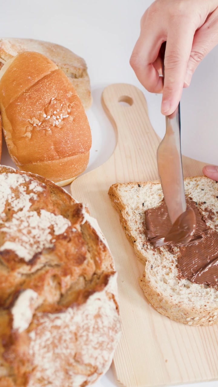 Hand spreads chocolate cream on bread slice amidst sourdough, baguettes, and croissants on wooden board.