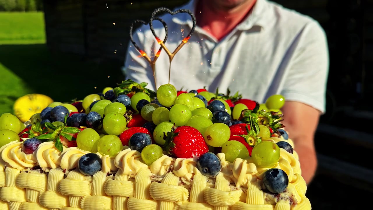 Uomo irriconoscibile che tiene in mano una torta di frutta decorata con frutti di bosco, uva, decorazioni di panna e un topper a forma di cuore scintillante, nella calda luce serale esterna.