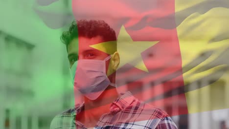 Animation-of-flag-of-cameroon-waving-over-latin-man-wearing-face-mask-in-city-street