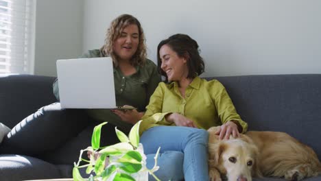 Caucasian-lesbian-couple-using-laptop-and-sitting-on-couch-with-dog