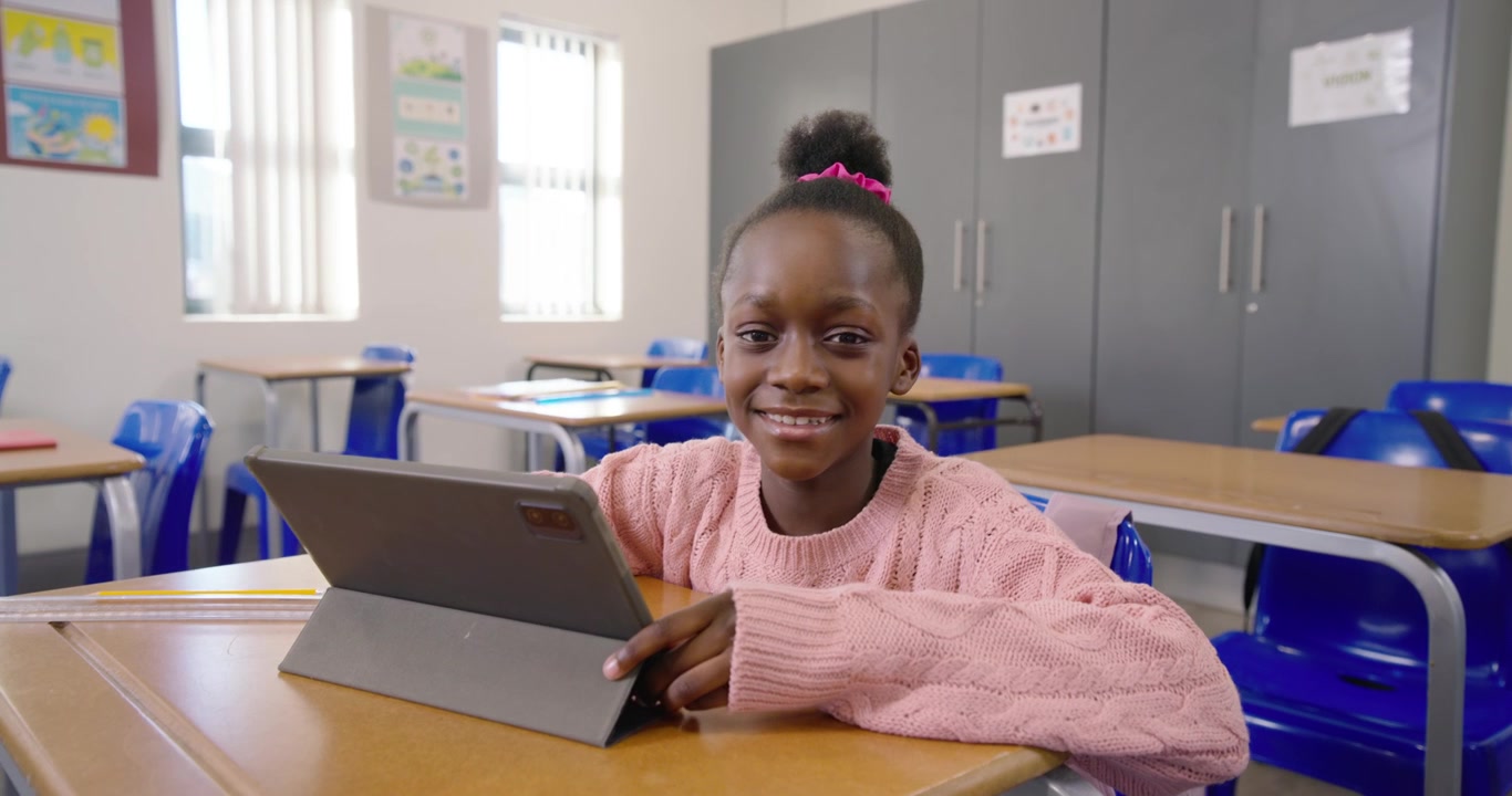 Bambino africano con maglione rosa che appoggia un tablet con custodia su un banco di scuola, toccando un'app sorridente e coinvolgente