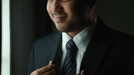 confident businessman in classic blue suit tying or adjust necktie near window in hotel room at the morning. handsome man wearing a nice suit on wedding day.