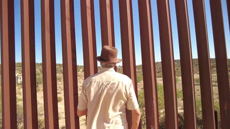 Focus on man with hat looking through US Mexico border fence, then reversing and showing barbed wire on top