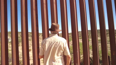 focus on man with hat looking through us mexico border fence, then reversing and showing barbed wire on top