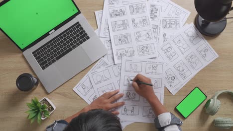 vista de cerca de arriba del artista masculino asiático dibujando el guión gráfico para la película en la mesa con pantalla verde teléfono inteligente y portátil en el estudio