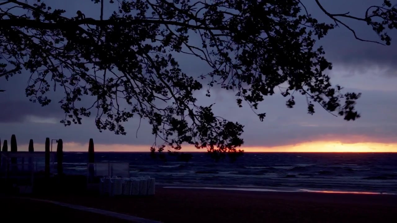 Static shot captures dramatic sunset glow on Baltic Sea horizon framed by silhouetted tree branches and empty beach furniture in Jurmala during peaceful summer dusk.