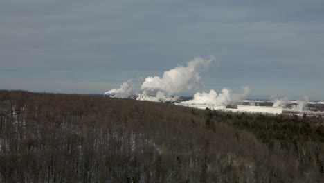 Flying-Over-Bare-Trees-In-Forest-With-Industrial-Plant-Of-Paper-Mill-Factory-Producing-Thick-Smoke-In-Quebec,-Canada