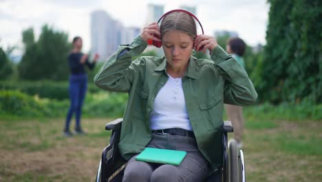 disabled caucasian girl in wheelchair putting on headphones opening tablet in slow motion. friends playing ball at background. portrait of sad abandoned teenager in summer park outdoors.