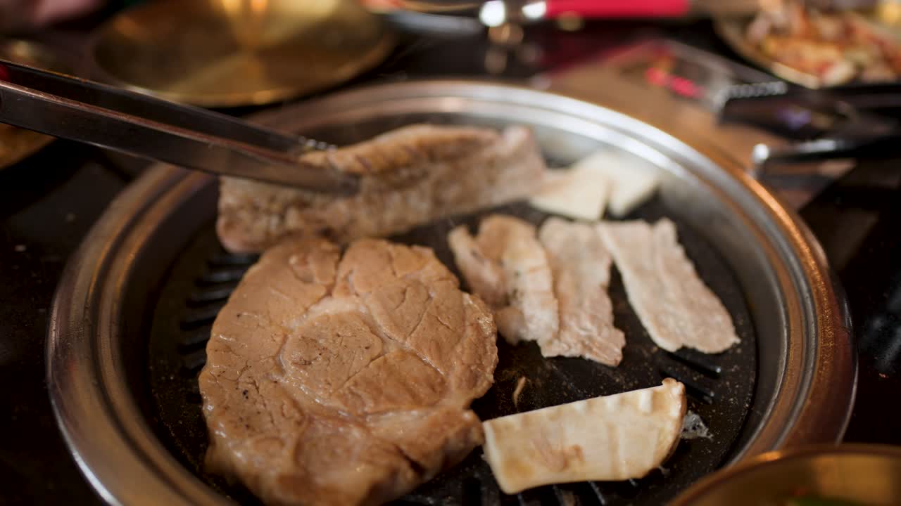Hand uses tongs to flip pork belly slices on tabletop Korean barbecue grill, warm lighting