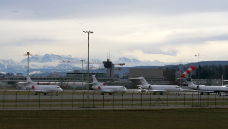 a line up of big corporate jets at an airport, white snowy mountains in the background.