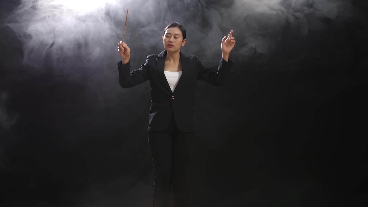 안개가 있는 검은 스튜디오에서 눈을 감고 몸짓을 하는 아시아 여성 지휘자