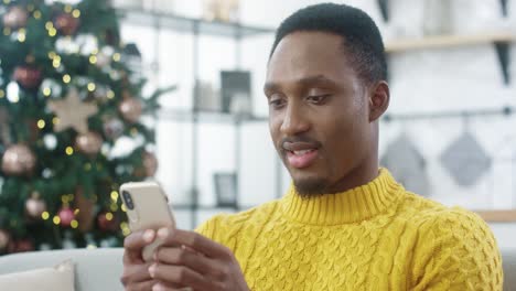 Primer-Plano-Retrato-De-Hombre-Alegre-En-Suéter-Amarillo-Sentado-En-Casa-Con-árbol-De-Navidad-Brillante-Y-Mensajes-De-Texto-En-El-Teléfono-Inteligente-Enviando-Saludos-Y-Deseos-A-Amigos-Feliz-Año-Nuevo-Concepto