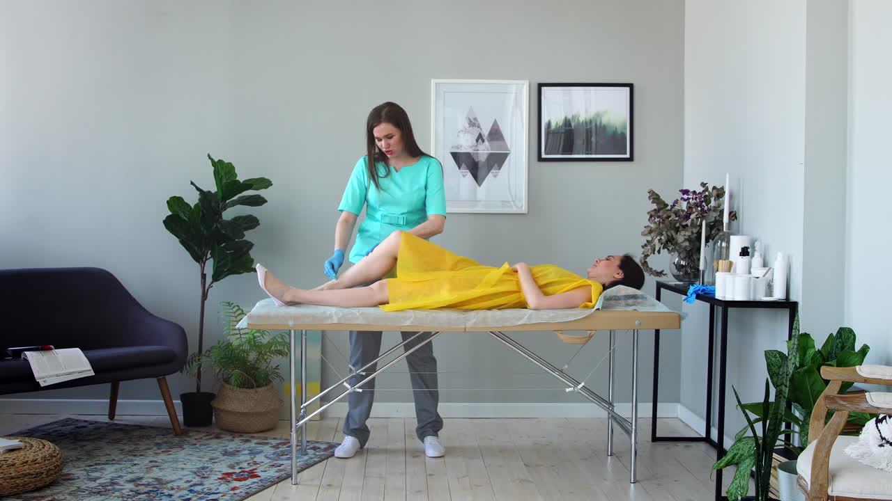 dos chicas en un salón de belleza maestro en una bata y guantes haciendo el procedimiento para eliminar el pelo en las piernas con una mezcla de azúcar