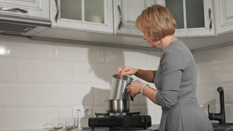 housewife cooking stirring soup inside metal jug placed inside pot on gas burner while arranging glass cups with candle wicks nearby on kitchen countertop, showing electric outlet on tiled wall