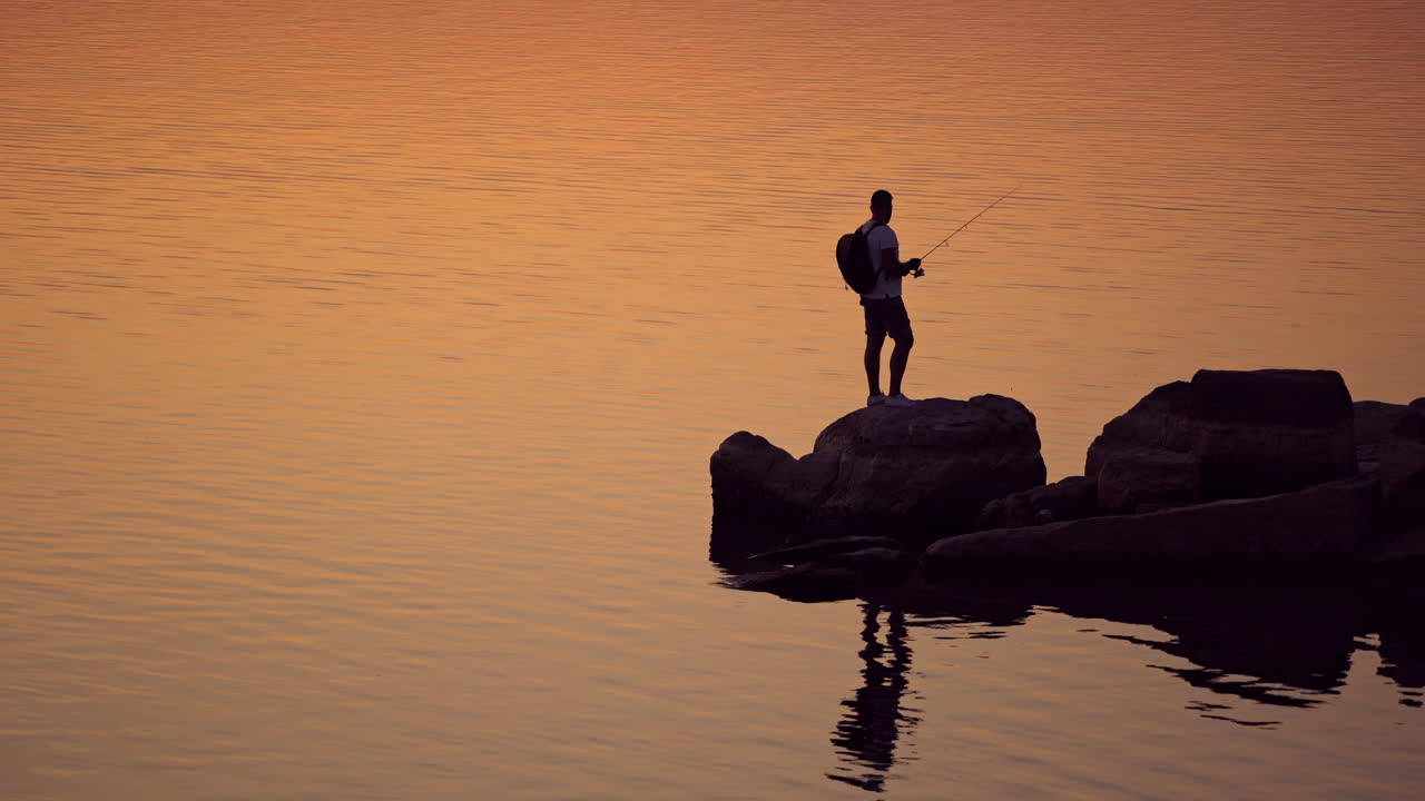 Silhouette of a fisherman fishing on the rock at sunset. Young man with rucksack is standing with fishing rod on a big stone near the river in the evening.