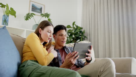 happy diverse couple sitting on sofa using tablet at home, in slow motion