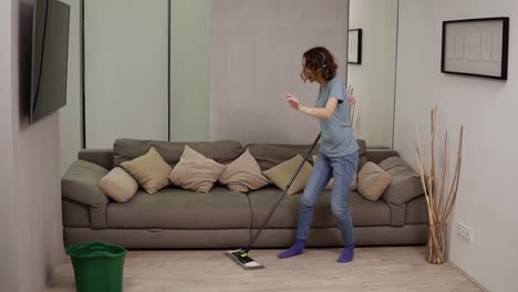 People,-housework-and-housekeeping-concept---happy-woman-in-headphones-with-mop-cleaning-floor-and-dancing-at-home.-Grey-couch-on-the-background.-Slow-motion