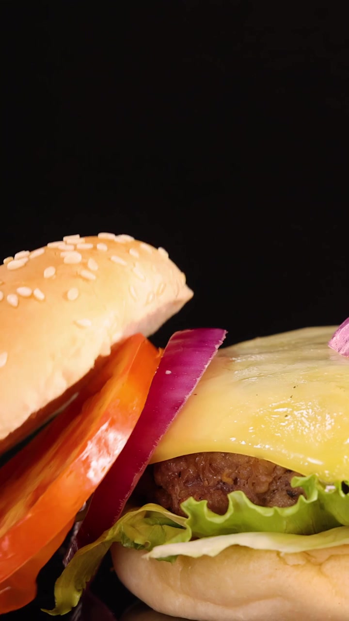 Un pan de sésamo y una rodaja de tomate caen sobre una hamburguesa con queso de carne de res con lechuga y cebolla, capturado en cámara lenta sobre un fondo negro con iluminación de estudio