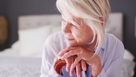 Happy-senior-caucasian-woman-sitting-on-bed-in-bedroom,-holding-walking-cane