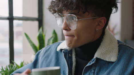 stylish young hispanic man student drinking coffee at home looking out window thinking planning ahead enjoying relaxing lifestyle wearing trendy fashion glasses