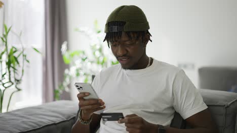 Young-black-man-holds-plastic-bank-card,-sitting-on-a-comfortable-sofa,-shopping-using-smartphone