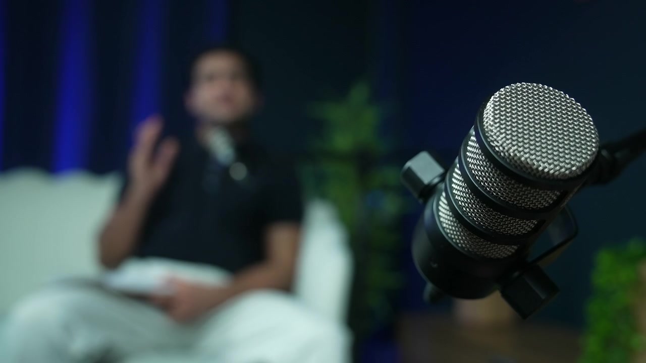 Ekstremt nærbillede af en dynamisk studiemikrofon med en sløret mandlig taler i baggrunden. Perfekt til podcasting, broadcasting og koncepter inden for lydproduktion.