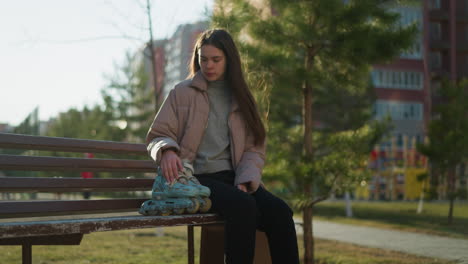 a young girl sits on a park bench, wearing a peach jacket, grey inner shirt, and black trousers. her rollerblades rest beside her on the bench as the wind gently blows her hair