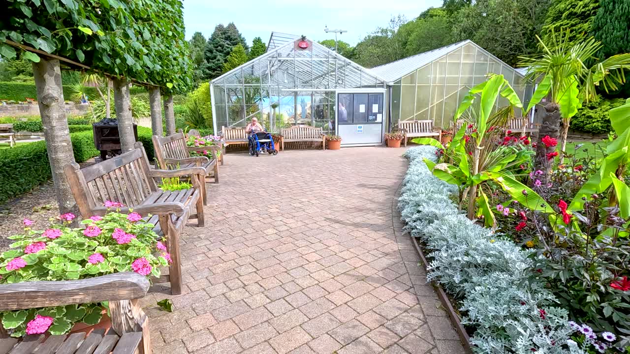 A steady daytime walk along a brick path lined with benches and vibrant flowerbeds, approaching a greenhouse in a lush botanical garden