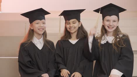 happy kindergarten students in cap and gown smiling and looking at the camera while sitting on stairs during the preschool graduation ceremony