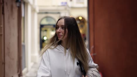 A-woman-walks-along-a-city-narrow-street-with-backpack,-looks-with-interest-around