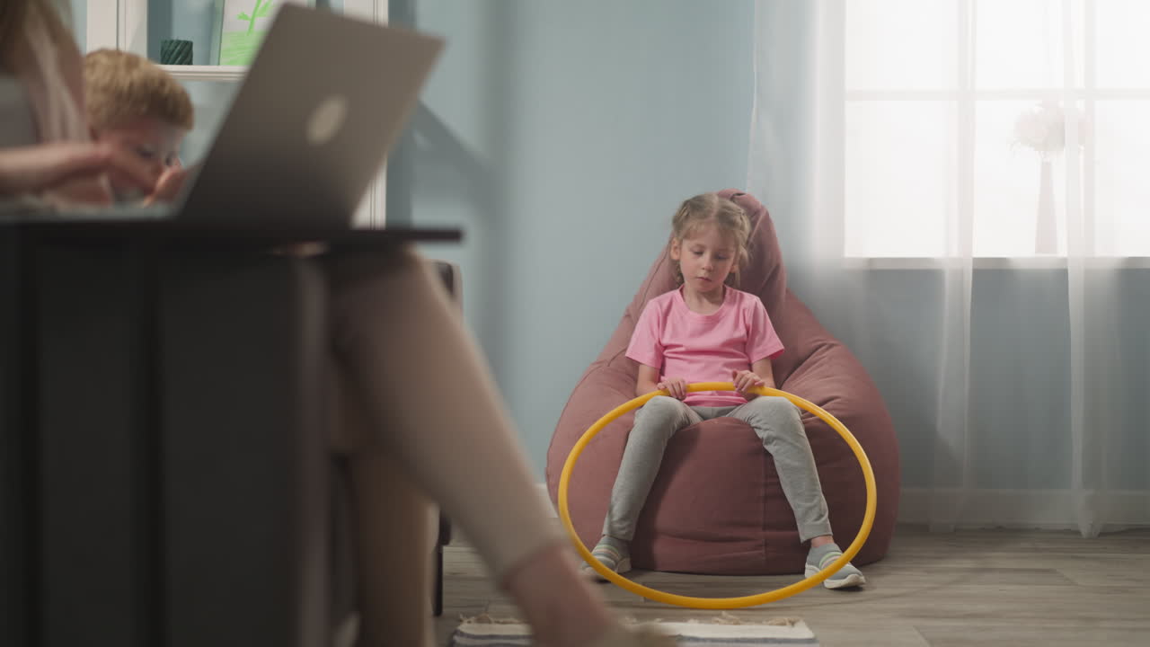 Sad little girl sits in bean chair mother works on laptop