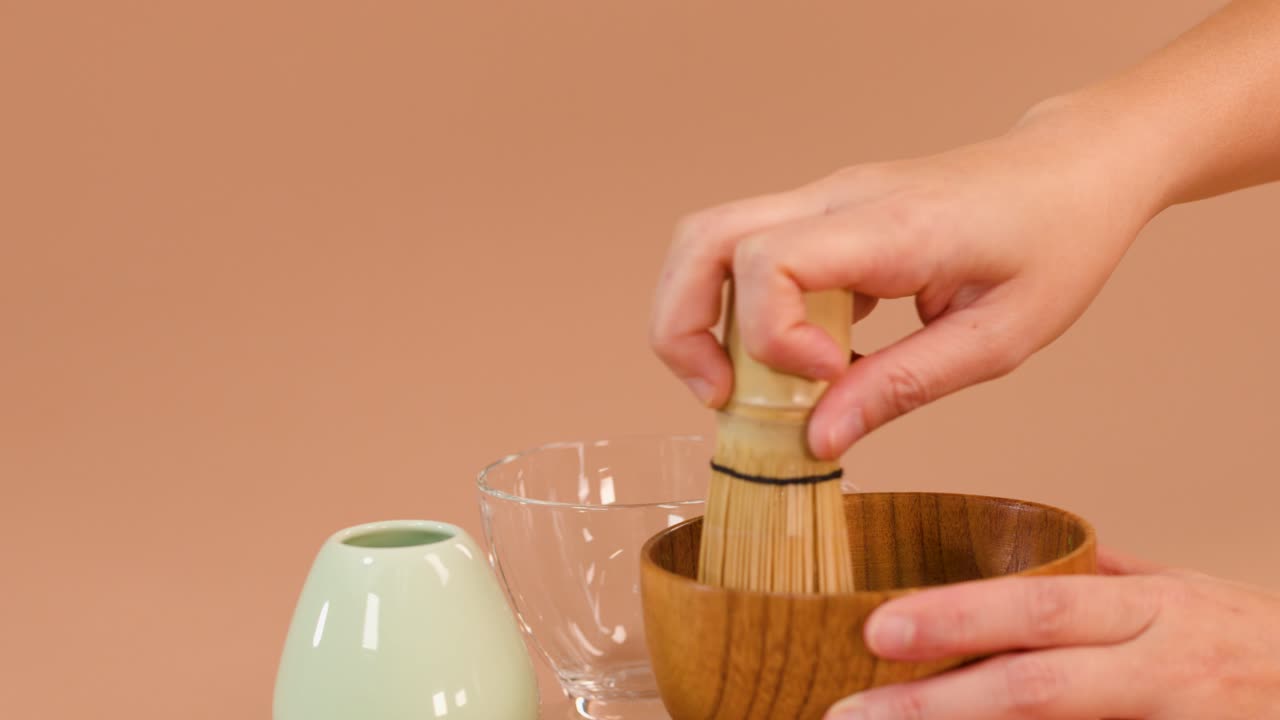 Hands whisk matcha powder in wooden bowl with bamboo whisk, soft lighting, neutral background