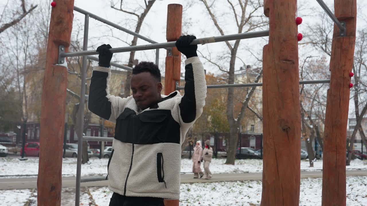 niño negro en una chaqueta de invierno salta para agarrar una barra de hierro, realizando pull-ups en un parque urbano nevado con un fondo de árboles desnudos y transeúntes cerca de los edificios