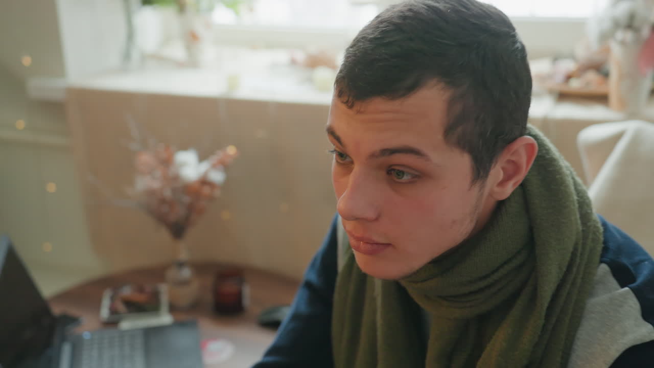 Uomo bianco con sciarpa che studia al laptop. Scena domestica tranquilla mostra studente e scrittore che studiano vicino alla finestra con fiori secchi, calde luci bokeh, primi piani spontanei dai toni tenui catturano la concentrazione.